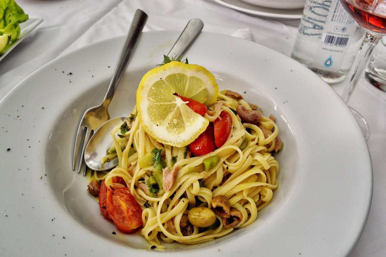 Piatto di bigoli integrali freddi con zucchine a julienne verde brillante, pomodori rossi, carote arancioni, basilico fresco e filo di olio extravergine, servito in ciotola bianca su sfondo beige con 