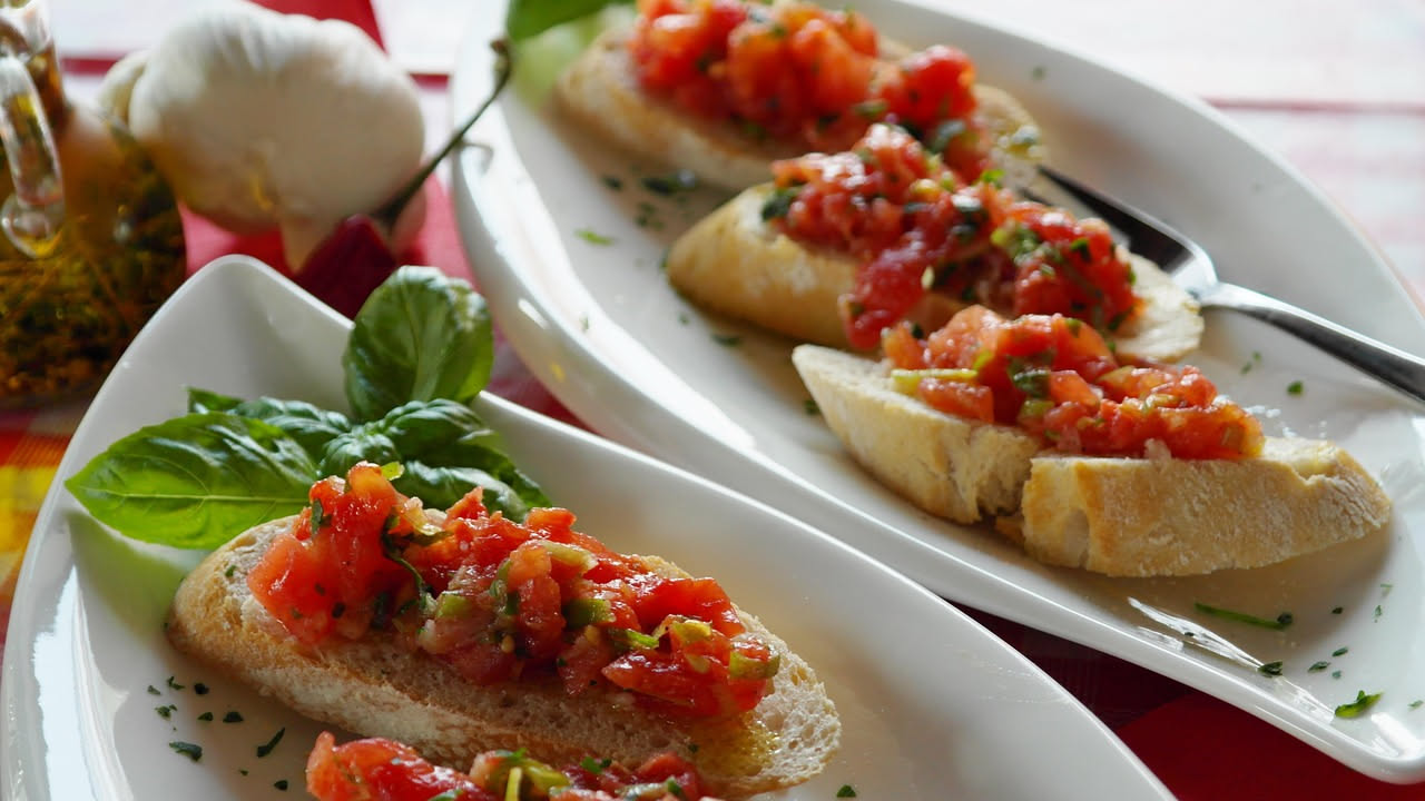 Tre fette di pane tostato croccante coperte di pomodori rossi a dadini, basilico verde fresco e gocce d'olio d'oliva, disposte su un piatto bianco con accanto un tovagliolo di lino