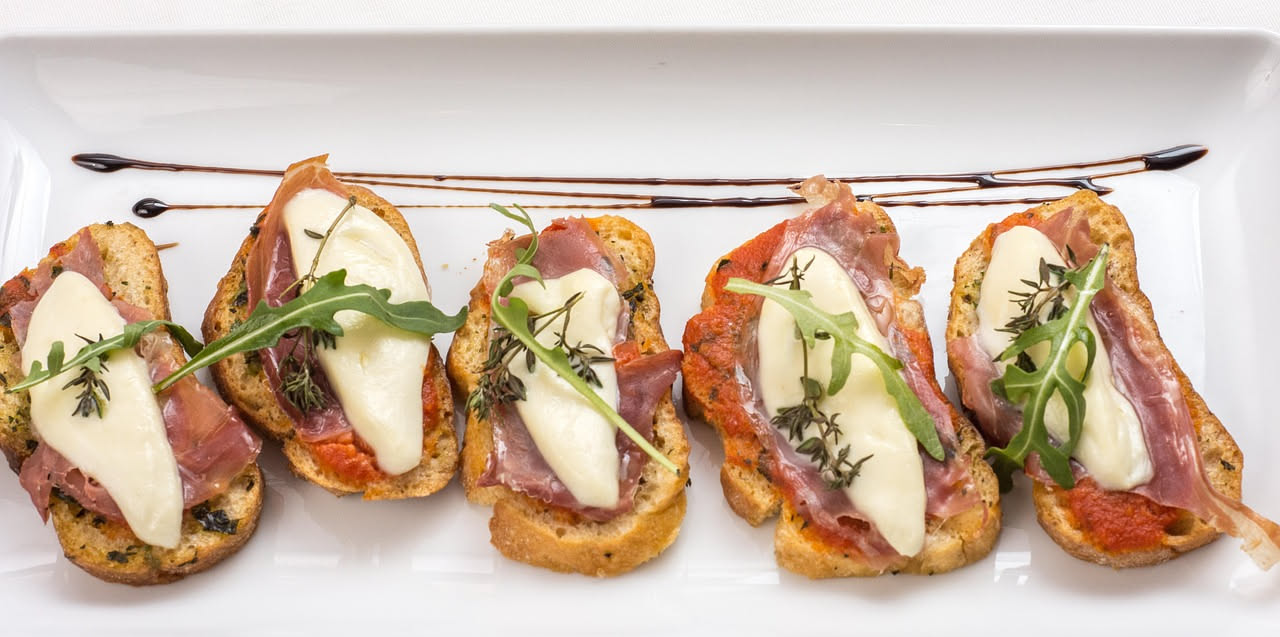 Sei bruschette su piatto bianco con toppings diversi: una con pomodori e basilico, una con ricotta e rucola, una con olive e aglio, una con melanzane, una con tonno, una con zucchine grigliate. Pane d