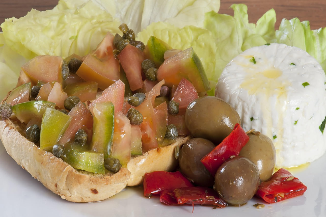 Caponata di melanzane in ciotola ceramica: melanzane brune frastagliate, dadi di pomodoro rosso, olive nere lucide, riflessi d'olio, coriandolo verde fresco, tavolo legno con luce naturale