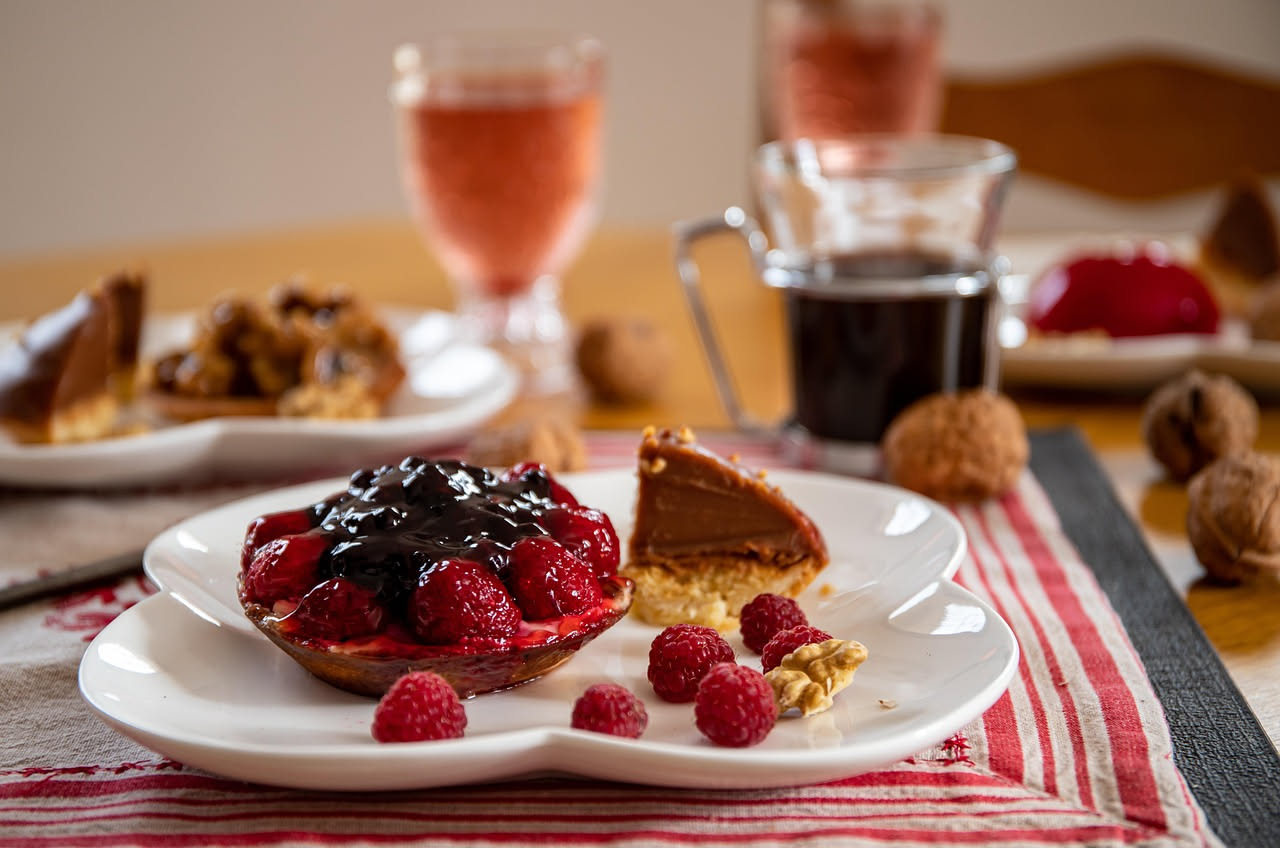 Crostata tonda su piatto bianco: guscio di pasta frolla dorata, crema gialla chiara visibile dentro, coronata di fragole rosse, mirtilli blu, lamponi rosa e more scure, spolverizzata di zucchero a vel