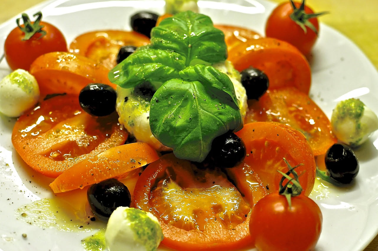 Piatto bianco con fette di pomodoro rosso, mozzarella di bufala bianca e foglie verdi di basilico fresco, condito con olio d'oliva