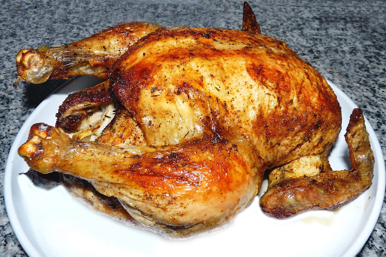 Pollo intero dorato e croccante appena sfornato, con limone e erbe aromatiche intorno, su un vassoio di presentazione bianco
