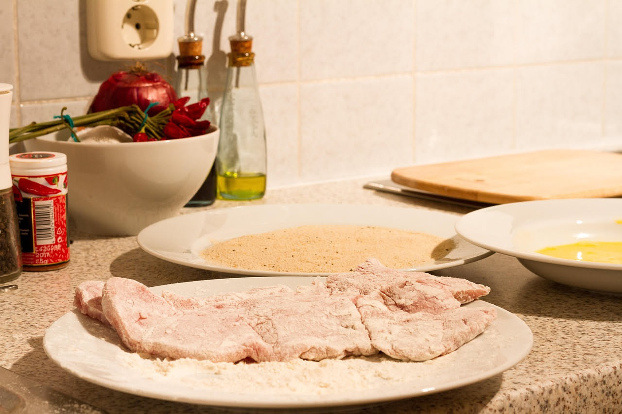Scaloppina di vitello coperta di farina bianca, accanto a una padella in acciaio e una salsa color ambrato in una ciotola bianca