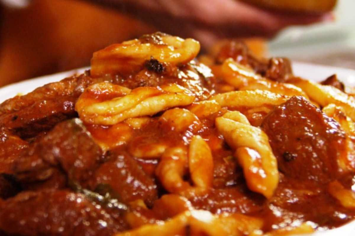 Pentola di terracotta con spezzatino di carne rosso intenso, pezzi di cipolla e carota, su fuoco acceso, vapore che sale dalla superficie