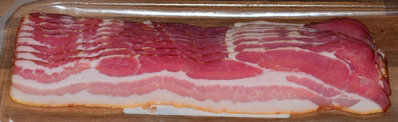 Pancetta di Sarconi affumicata, affettata in fette spesse su un tagliere di legno, con colorazione rosa dorato e striature di grasso bianco, fumo visibile sullo sfondo