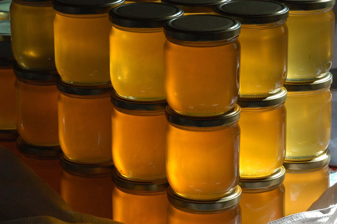 Barattolo di vetro trasparente contenente miele color ambra, uno più scuro e denso a sinistra, uno più chiaro e liquido a destra, su tavolo di legno con luce naturale estiva