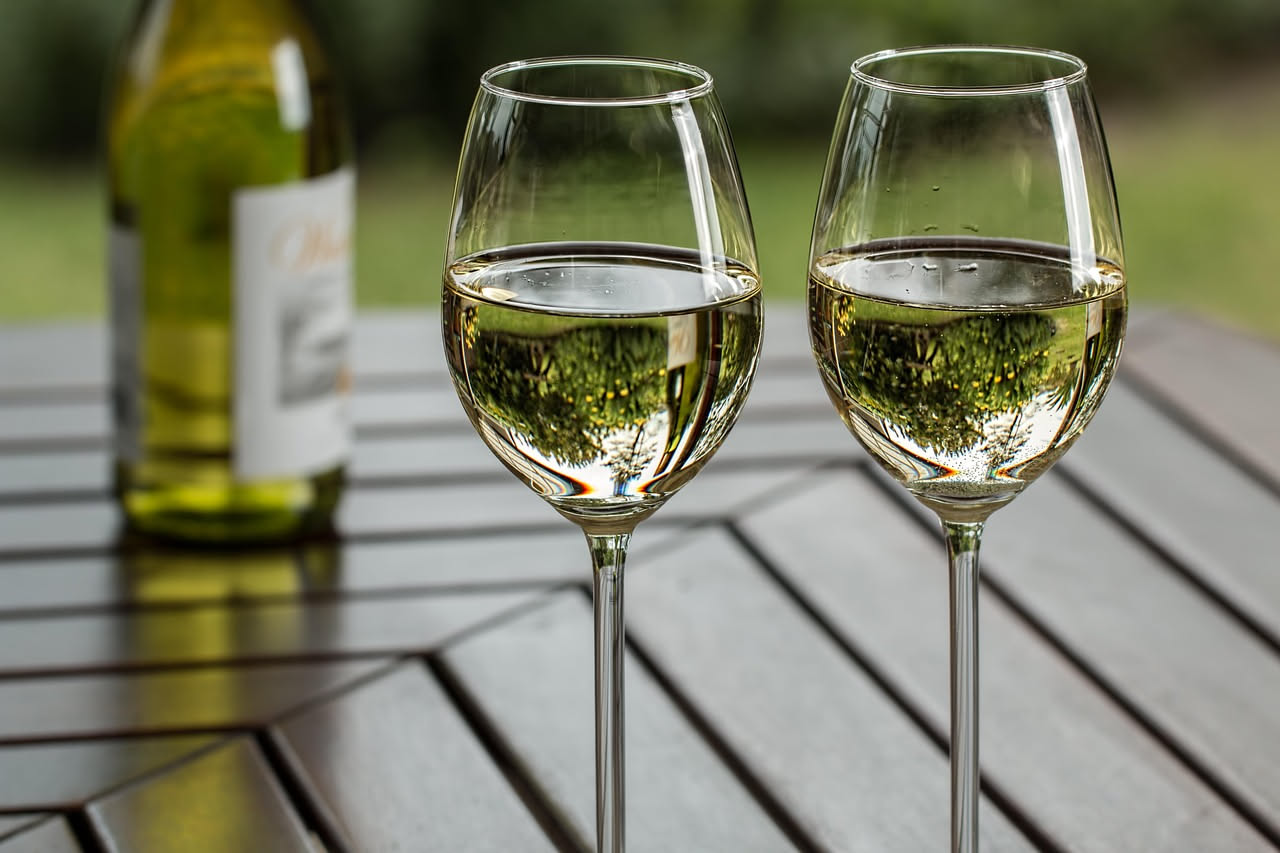 Calice di vino bianco ghiacciato con gocce di condensa, servito accanto a una bottiglia in secchiello con ghiaccio, su tavolo all'aperto estivo con sfondo di fogliame verde