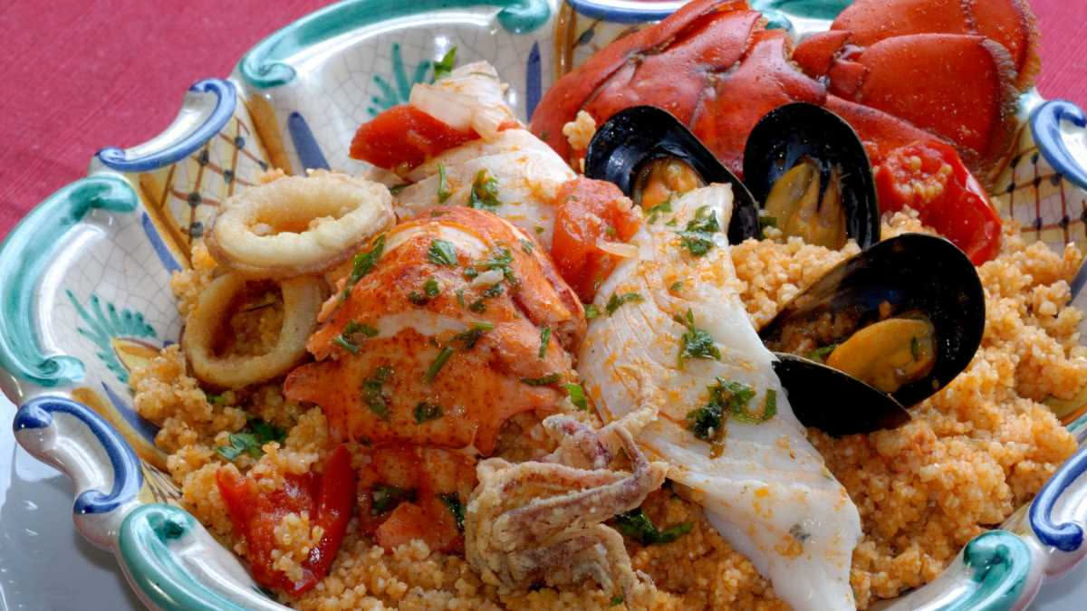 Cous cous di pesce trapanese in un piatto bianco con brodo di pesce, filetti di pesce azzurro, pomodori e cipolla: colori caldi ambrati e profumi di mare