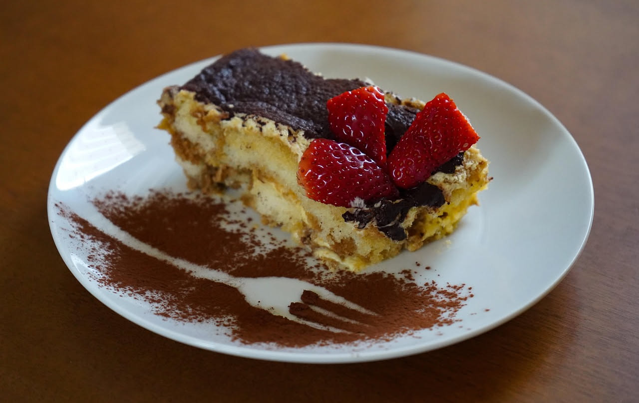 Tiramisù alle fragole in piatto bianco, strati visibili di crema mascarpone bianca e fragole rosse, guarnito con fragola fresca intera e qualche briciola di biscotto sulla superficie