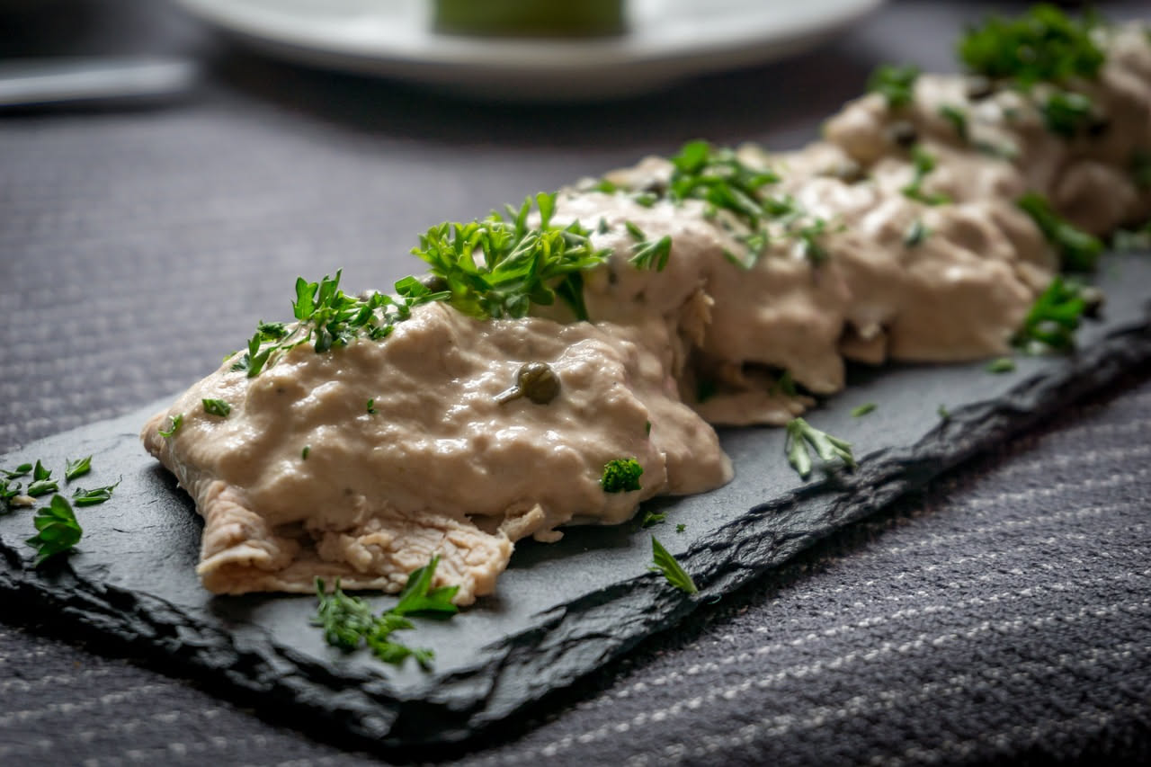 Piatto di vitello tonnato freddo con fettine di carne bianca ricoperte di salsa tonnata cremosa, decorate con limone sottile e capperi verdi su fondo bianco