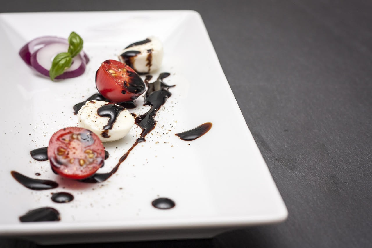 Piatto bianco con insalata caprese: fette di pomodoro rosso brillante alternate a dischi di mozzarella bianca, foglie di basilico verde fresco, gocce di olio d'oliva, sale e pepe nero visibili