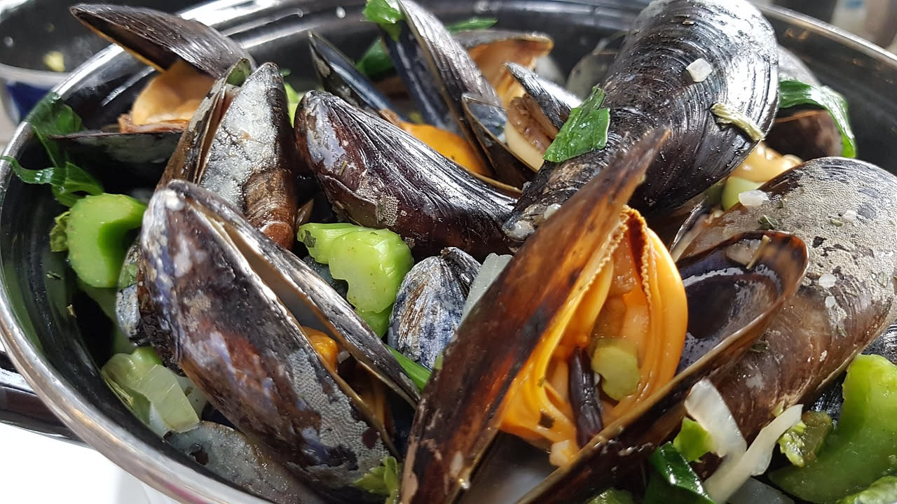 Cozze nere e vongole grigie su un tagliere di legno, con un coltello a lama corta, uno spazzolino bagnato e un recipiente d'acqua fredda sullo sfondo