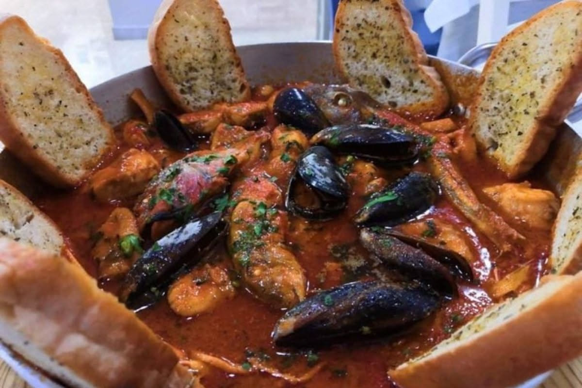 Padella in acciaio inox con gamberi rossi, calamari bianchi e cozze nere che saltano nel vapore su fuoco vivo, con aglio e prezzemolo fresco visibili