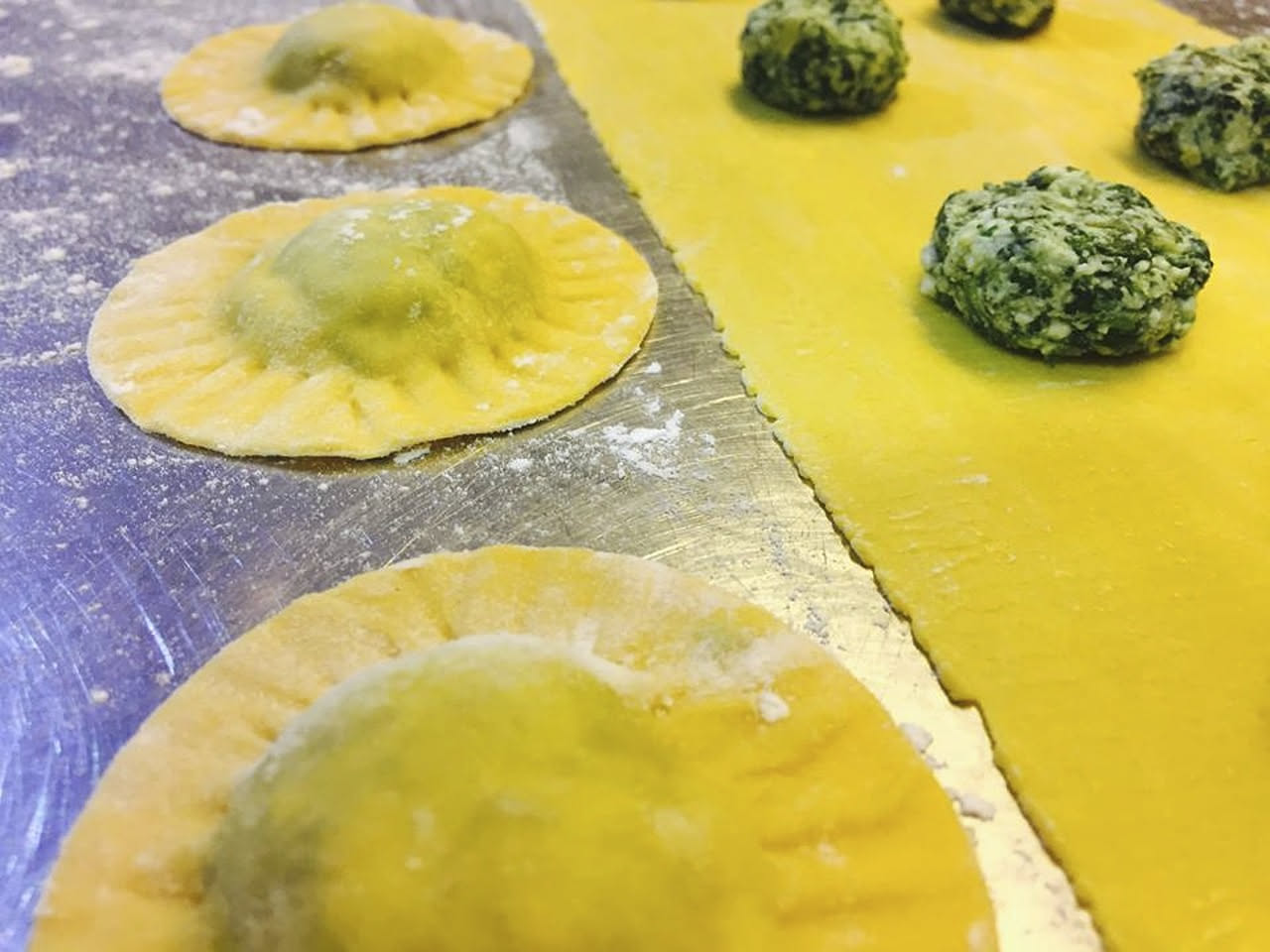 Ravioli appena chiusi su un tavolo di legno bianco, con una mano che sigilla un bordo con le dita leggermente infarinate, accanto un tegame di acqua bollente con vapore