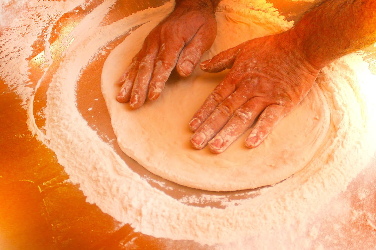 Impasto di pizza in una ciotola durante il riposo, con bolle di fermentazione visibili sulla superficie e colore dorato, accanto a un bicchiere d'acqua