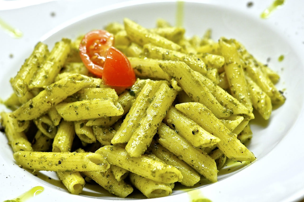 Piatto di pasta rigatoni condita con pesto verde intenso di rucola, pinoli tostati e lamelle di parmigiano grattugiato fresco, decorato con una foglia di rucola cruda