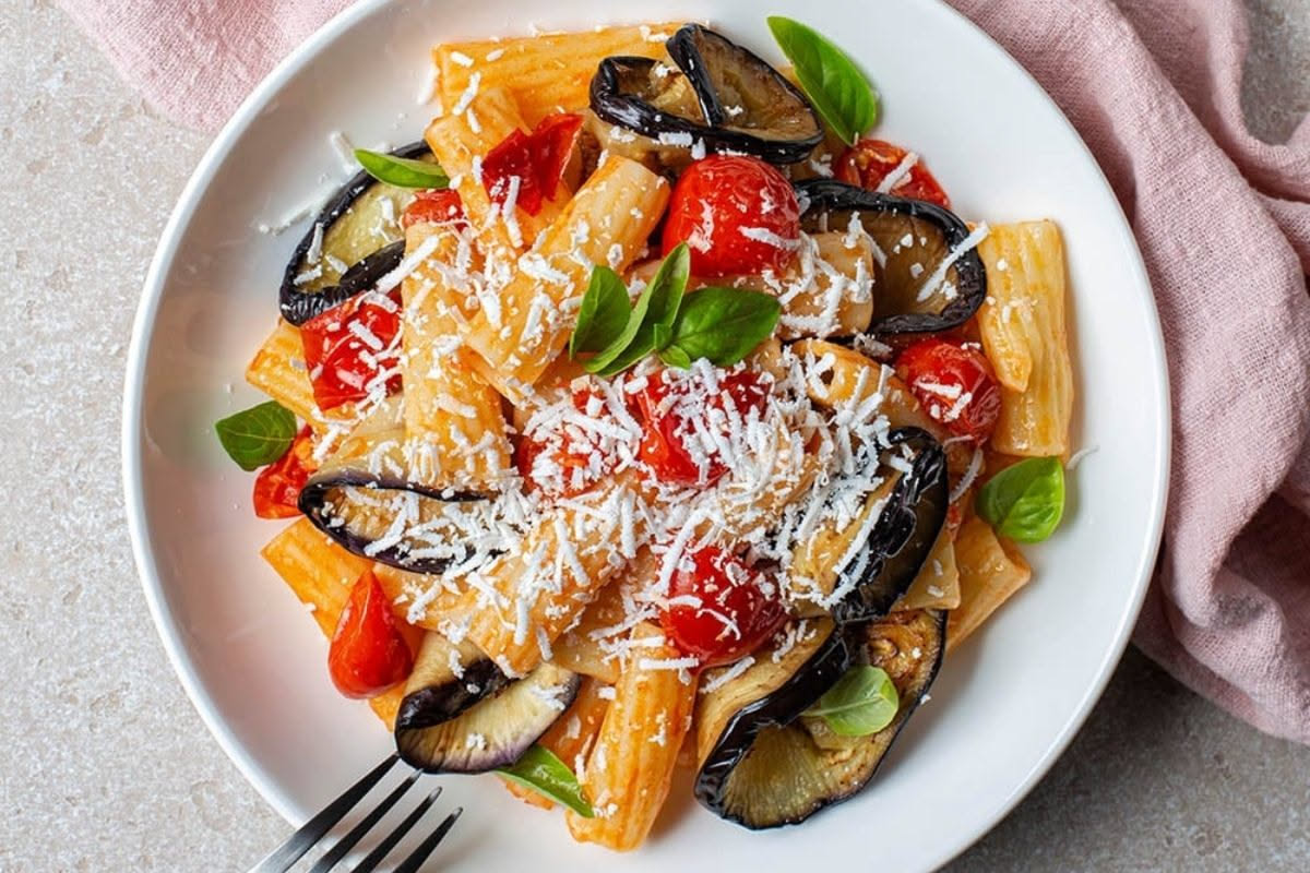 Piatto bianco con penne rigate condite con strisce di melanzana grigliata grigio-marrone, quadratini di mozzarella bianca filante, basilico fresco verde e salsa di pomodoro leggera, impiattate con uno