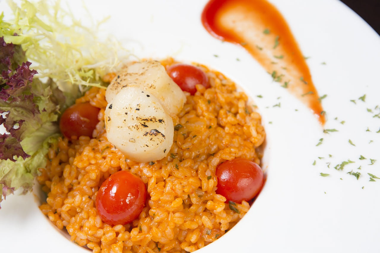 Piatto bianco con risotto cremoso dai toni rosati, pomodorini interi e spezzati fra il riso, parmigiano grattugiato in superficie, erba basilica fresca, sfondo sfocato di cucina
