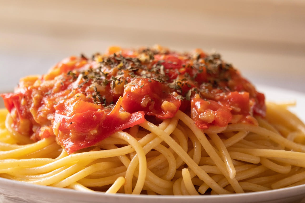 Piatto di spaghetti al dente conditi con pomodorini rossi tagliati a metà, foglie di basilico verde brillante, filo di olio d'oliva dorato, servito in ciotola bianca.