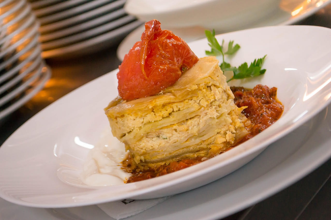 Tortino di melanzane in piatto bianco, dorato in superficie, con crosta di formaggio gratinato visibile, contorno di verdure fresche e salsa di pomodoro