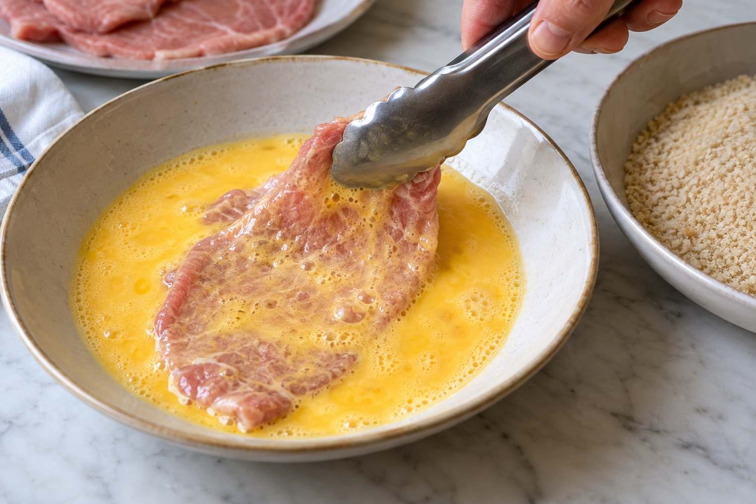 Una pinza immerge una fettina di carne nell'uovo sbattuto in un piatto fondo, accanto a una ciotola di pangrattato