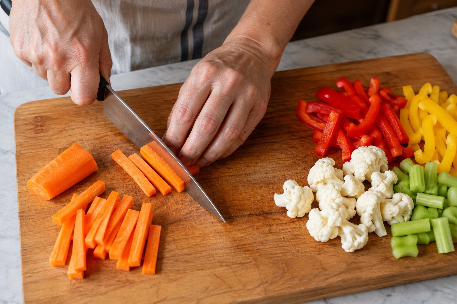 Una carota tagliata a bastoncini su un tagliere accanto a gruppi separati di peperoni, cavolfiore e sedano