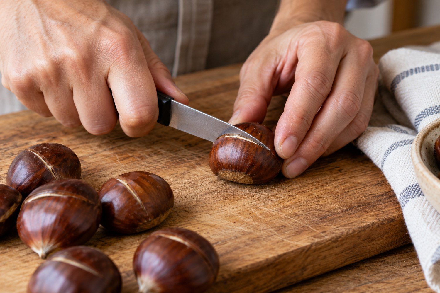 Una mano incide con un coltellino il lato bombato di una castagna appoggiata su un tagliere di legno, accanto ad altre castagne già incise