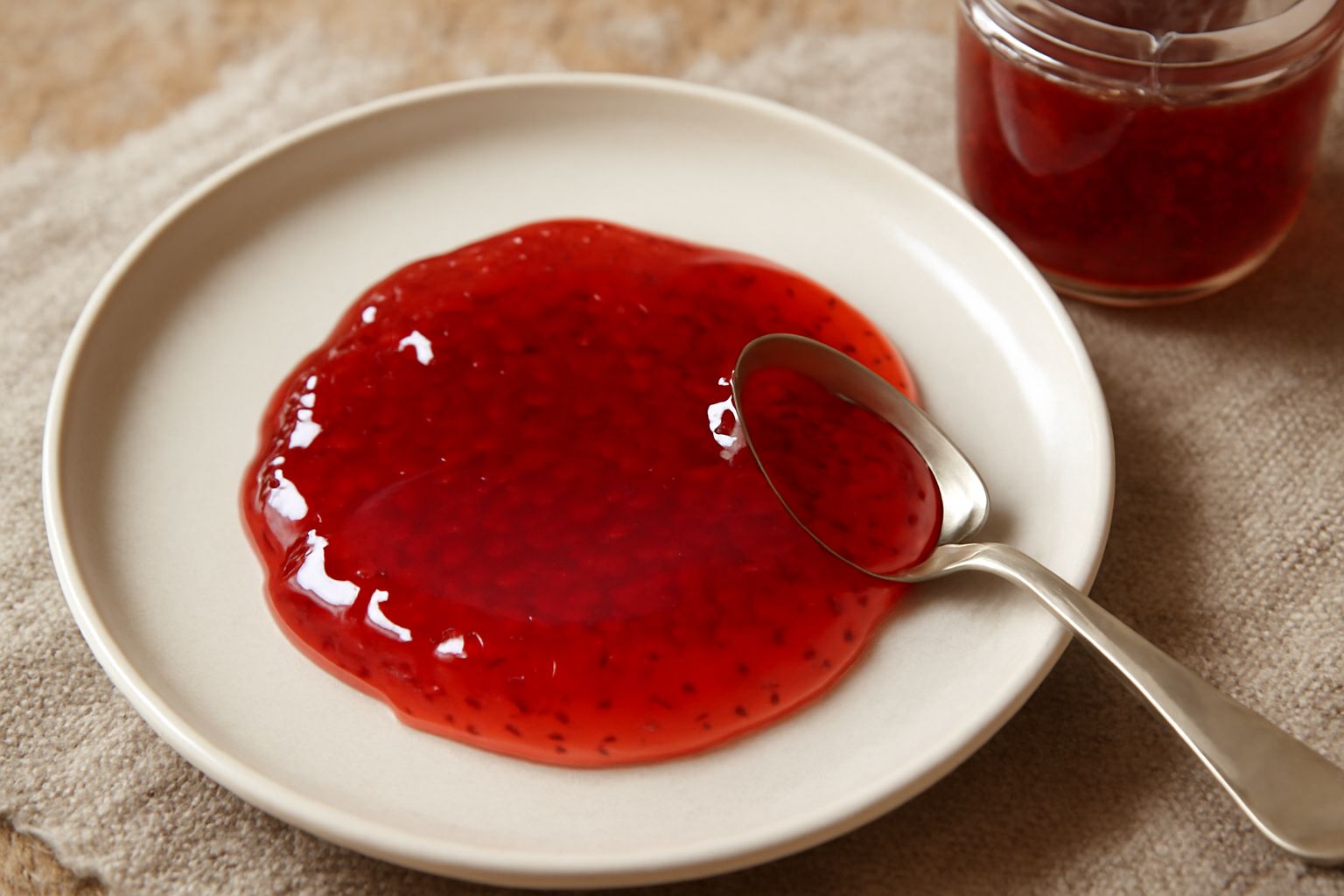 Vasetto di vetro con confettura rosso scuro e lucida di fragoline di bosco, completamente pieno, con cucchiaio di legno appoggiato sul bordo e sfondo bianco