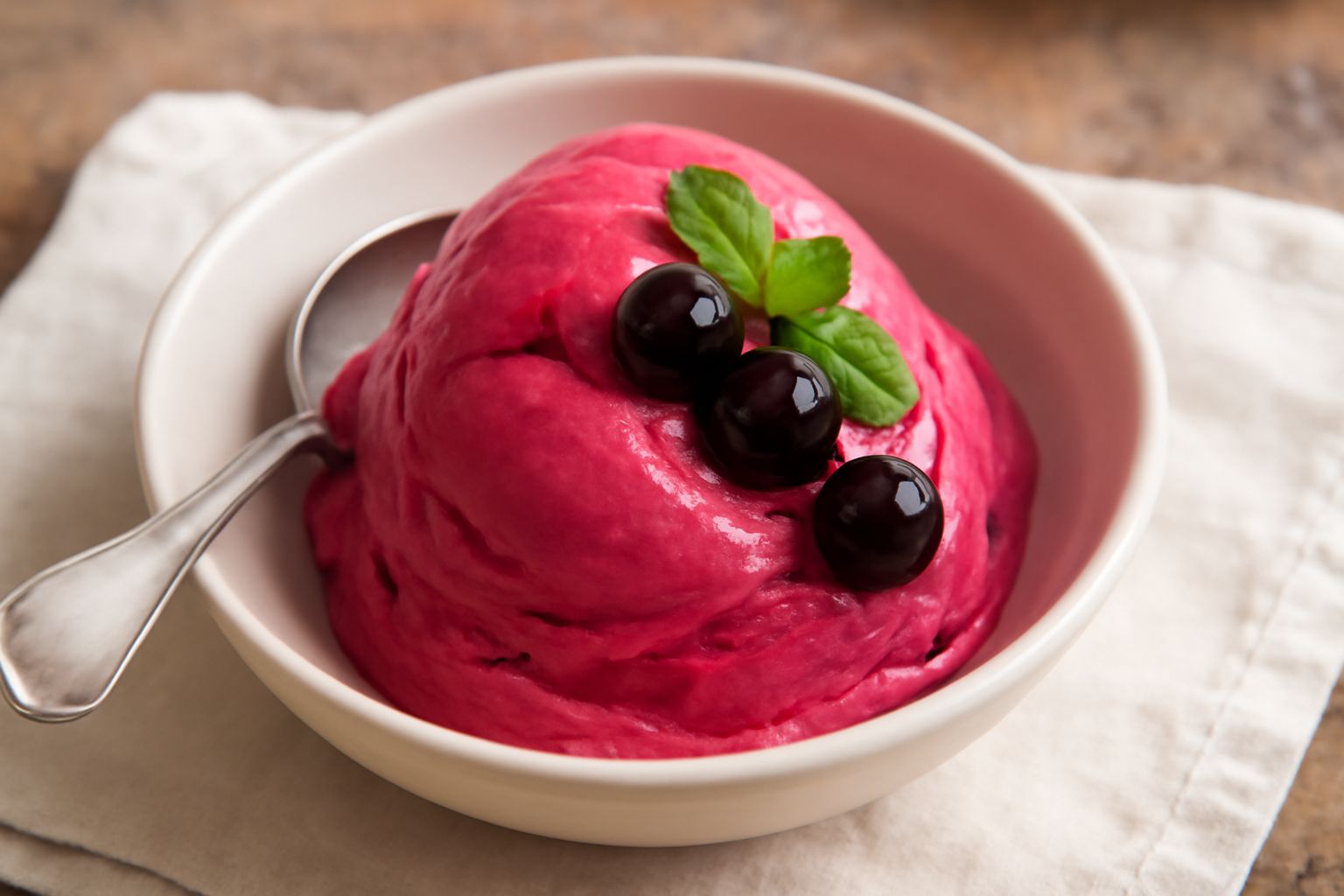 Gelato di amarena di colore rosa vivace in una coppetta di vetro, guarnito con un'amarena rossa e foglie di menta fresca, su un tavolo di marmo bianco con cucchiaio di legno