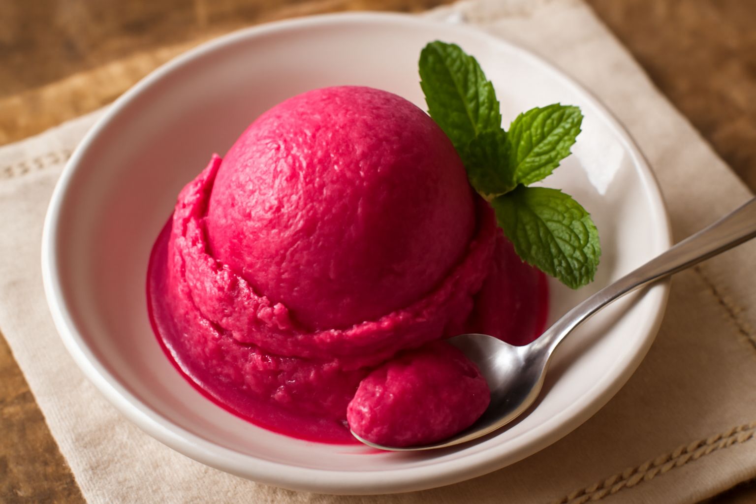 Gelato fico d'india rosa intenso servito in coppetta di vetro, con una pallina tonda e liscia, guarnito con una foglia di menta fresca e uno spicchio di fico d'india a lato