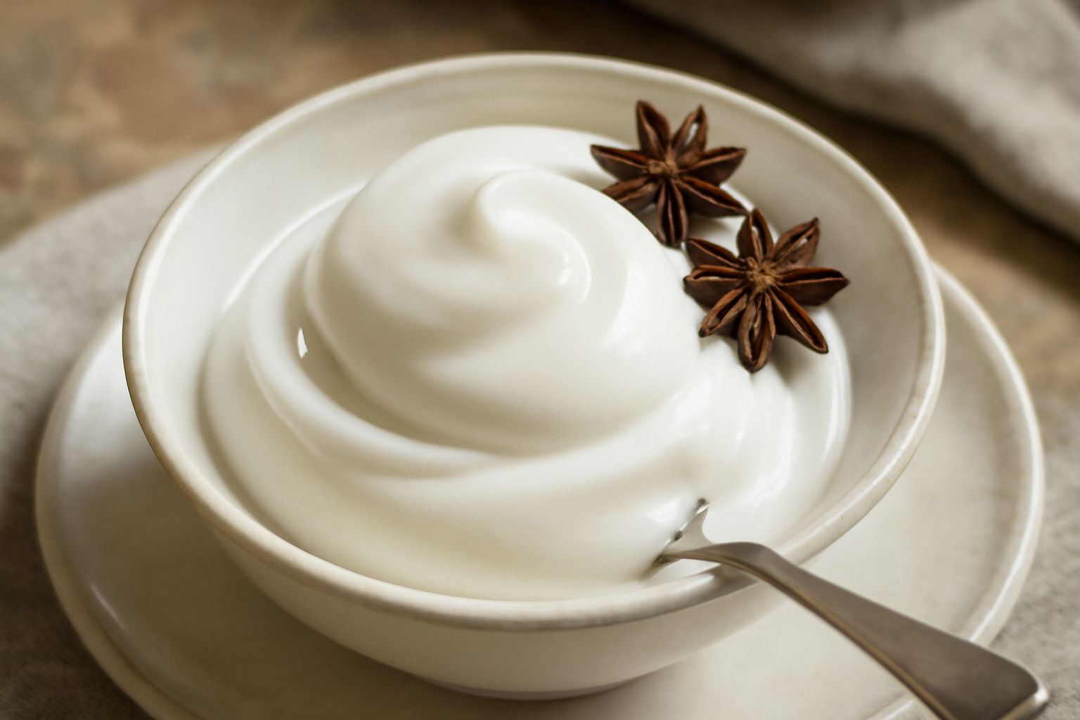 Gelato di sambuca di colore bianco panna, servito in coppetta di vetro con cucchiaio, superficie liscia e lucida, guarnizione di chicchi di anice stellato grigio sul bordo