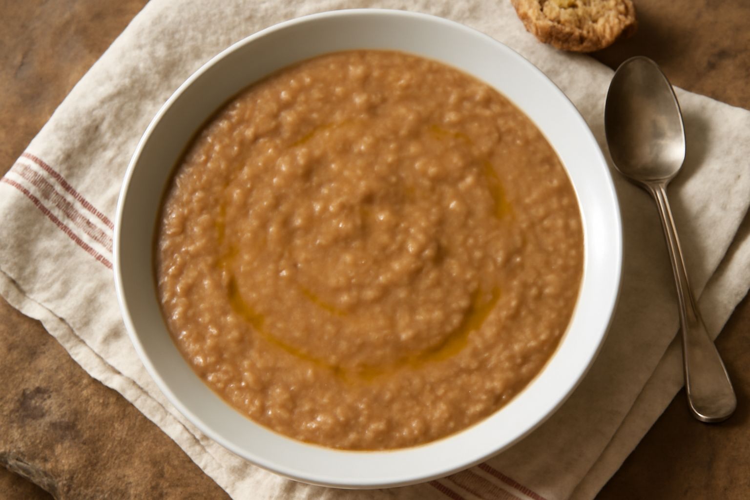Scodella di ceramica bianca con pappa di brodo beige-marrone, cremosa e omogenea, guarnita con un filo d'olio verde al centro e pezzettini di pane appena visibili, vapore leggero, cucchiaio di legno a