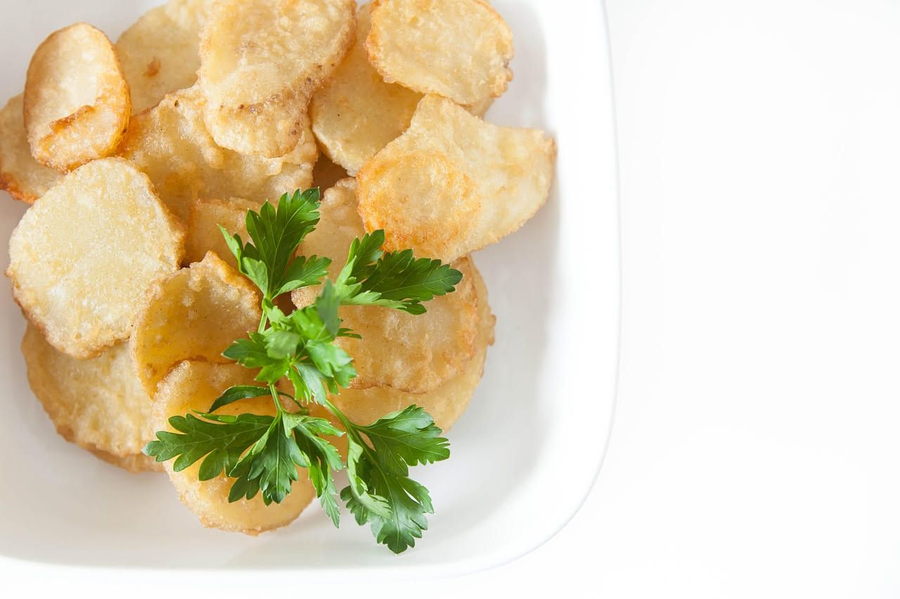 Patate chips dorate e croccanti in una ciotola di ceramica bianca, cosparse di sale marino grosso, con patate crude tagliate accanto e olio in una bottiglietta di vetro