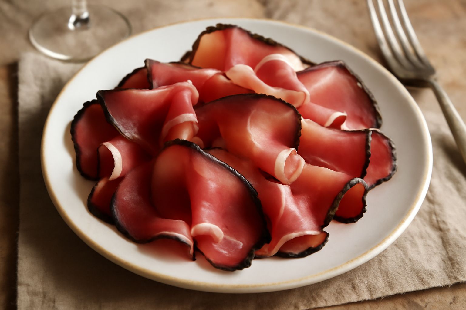 Speck dell'Oltrepò affettato sottile su un piatto bianco, con superficie rosa scuro e orli affumicati, disposto a ventaglio accanto a una fetta di pane tostato