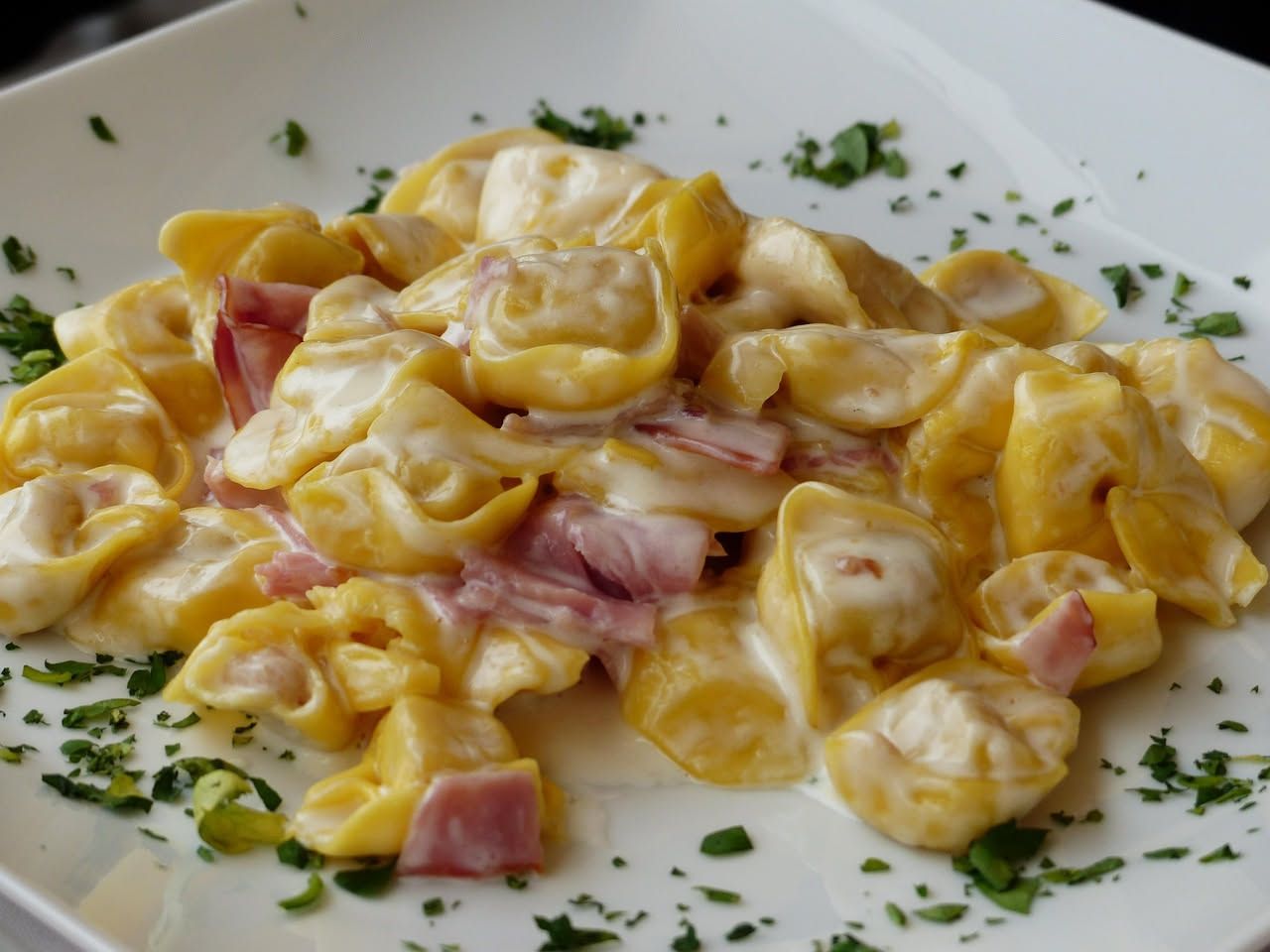 Tortellini freddi in una ciotola bianca con pomodori ciliegia, basilico fresco, olive nere e gocce di olio extravergine di oliva, su sfondo di lino grigio