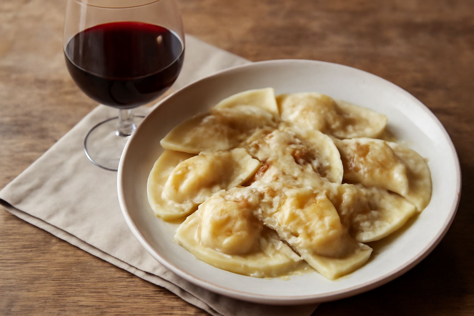 Bicchiere di vino rosso scuro Sassella della Valtellina accanto a un piatto di casunziei ripieni con patate, burro e salvia fresca su fondo di tavolo in legno naturale