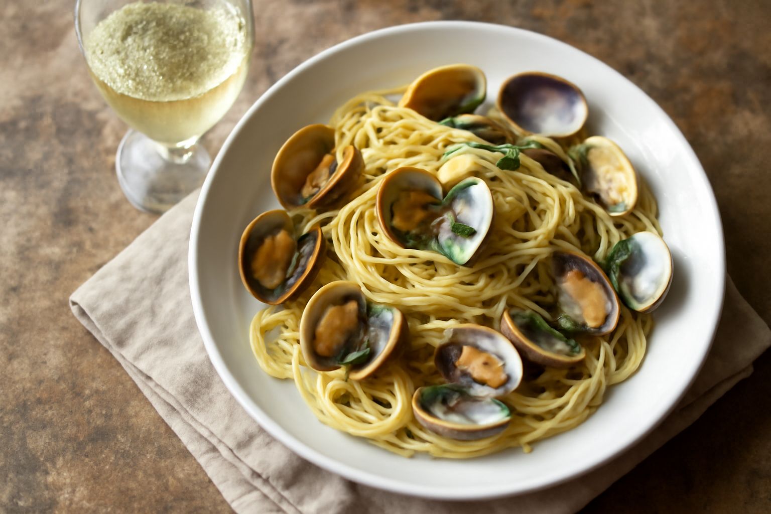 Bicchiere di vino bianco paglierino con riflessi dorati, calice in vetro cristallo su tavolo di legno naturale, accanto a piatto con branzino al forno, limone fresco e erbe aromatiche verdi.